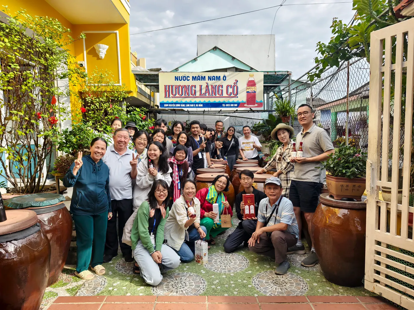 Nhóm người tụ tập bên nhau trước cửa hàng nước mắm Nam Ô với các chum lớn và bảng hiệu Hương Làng Cổ.