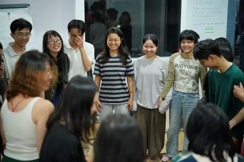 Nhóm thanh niên đang đứng và cười trong một phòng học với bảng viết chữ phía sau.